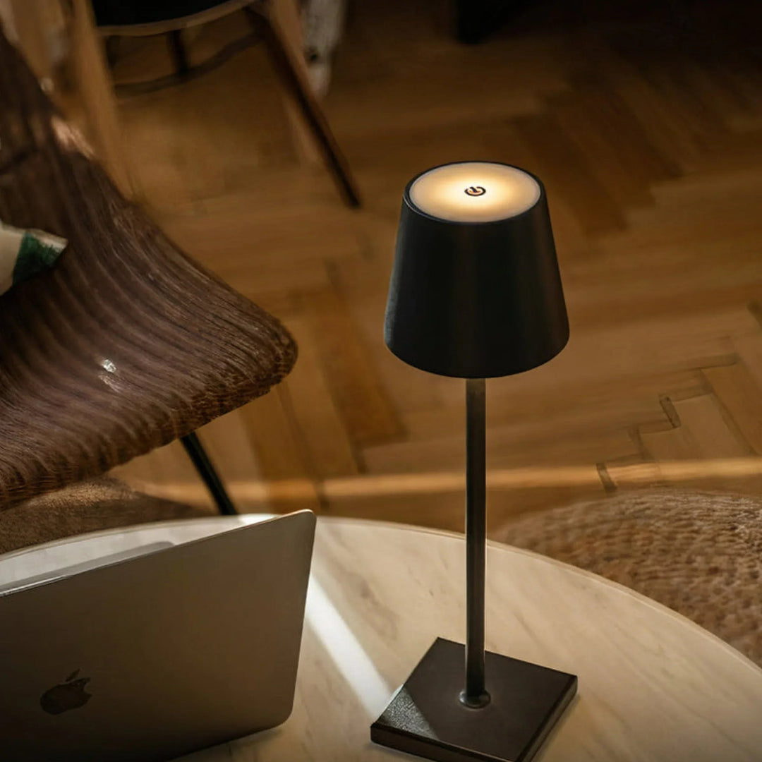Small black lamp on a table next to a laptop with a wooden chair in the background.